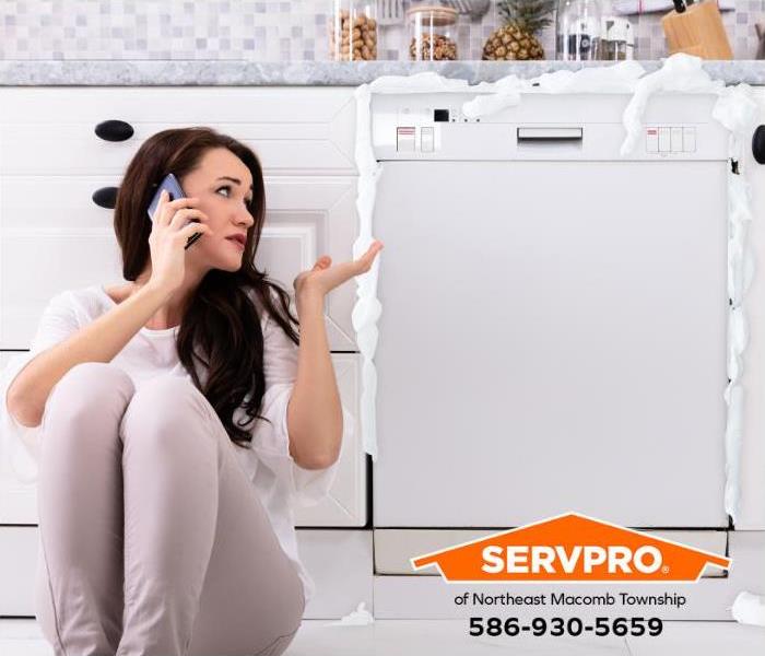 A person sits on the floor next to a dishwasher leaking soap suds while calling for help.